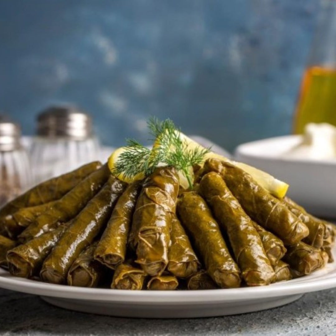 Grape leaves stuffed with seasoned rice, garnished with lemon wedges and fresh dill, are served on a plate. Salt and pepper shakers appear blurred in the background.