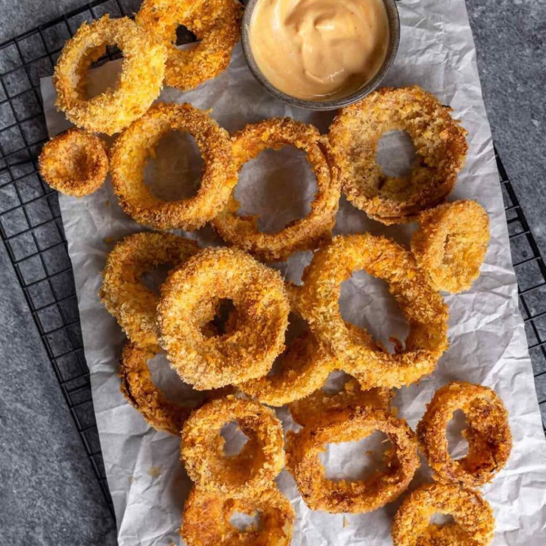 Onion Rings—golden and crispy with a crunchy breading—are served fresh from the fryer on parchment paper, paired with creamy dipping sauce, and set on a black wire rack over a gray surface.