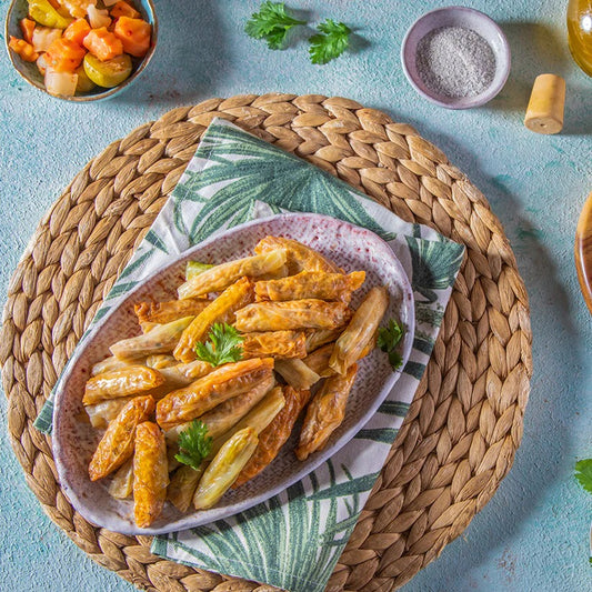 On a woven placemat, an oval plate of cabbage rolls filled with seasoned rice and garnished with parsley sits atop a green patterned napkin. Nearby are pickled vegetables, a dish of salt, and a glass of yellow beverage. Product: Cabbage.
