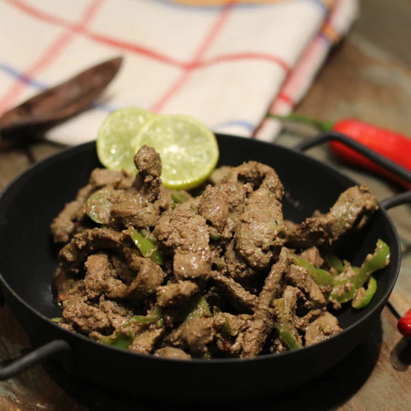 A black bowl holds Alexandrian Liver, a spicy stir-fried dish with sliced beef liver and green bell peppers, garnished with two lime slices. In the background are a cloth napkin, red chili, and cutlery on a rustic wooden surface.