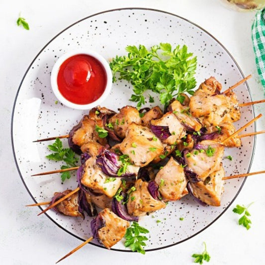 Shish Tawook chicken skewers with red onions are served on a white speckled plate, garnished with parsley. A small dish of ketchup and fresh herbs add the finishing touch to this vibrant grilled meal.