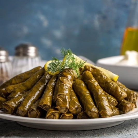 Grape leaves stuffed with seasoned rice, garnished with lemon wedges and fresh dill, are served on a plate. Salt and pepper shakers appear blurred in the background.
