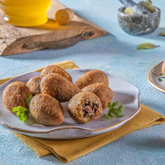 A plate of Kobiba, a Middle Eastern dish of bulgur and minced meat, is garnished with parsley. One Kobiba is cut open to reveal the savory filling. A yellow napkin and a wooden board with a bottle appear in the background.