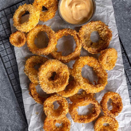 Onion Rings—golden and crispy with a crunchy breading—are served fresh from the fryer on parchment paper, paired with creamy dipping sauce, and set on a black wire rack over a gray surface.
