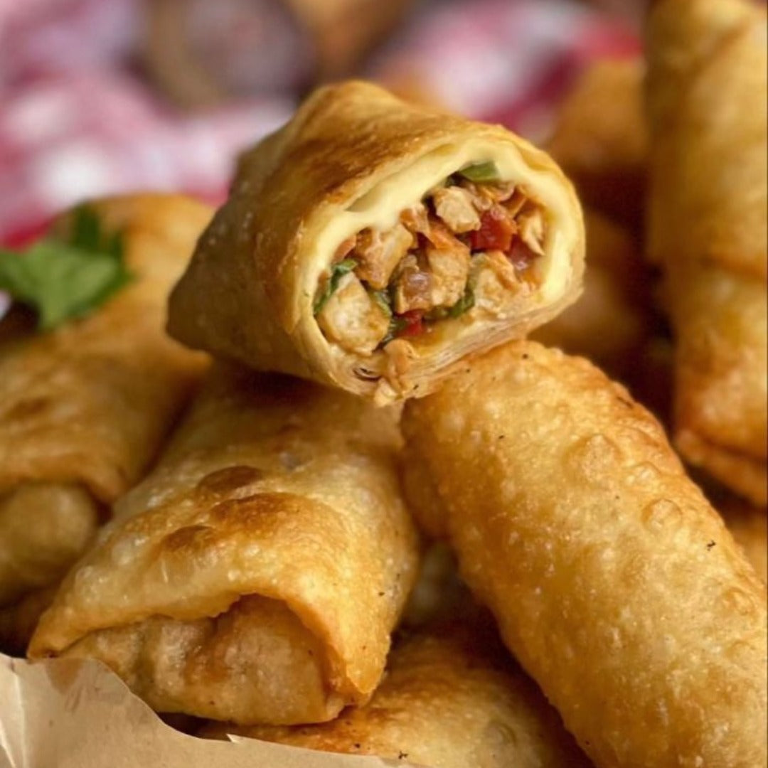 A close-up of crispy, golden-brown Spring rolls chicken, cooked in an air fryer, with one cut open to reveal a savory filling of chicken, veggies, herbs, and gooey mozzarella cheese. More Spring rolls chicken and a checkered cloth appear blurred in the background.