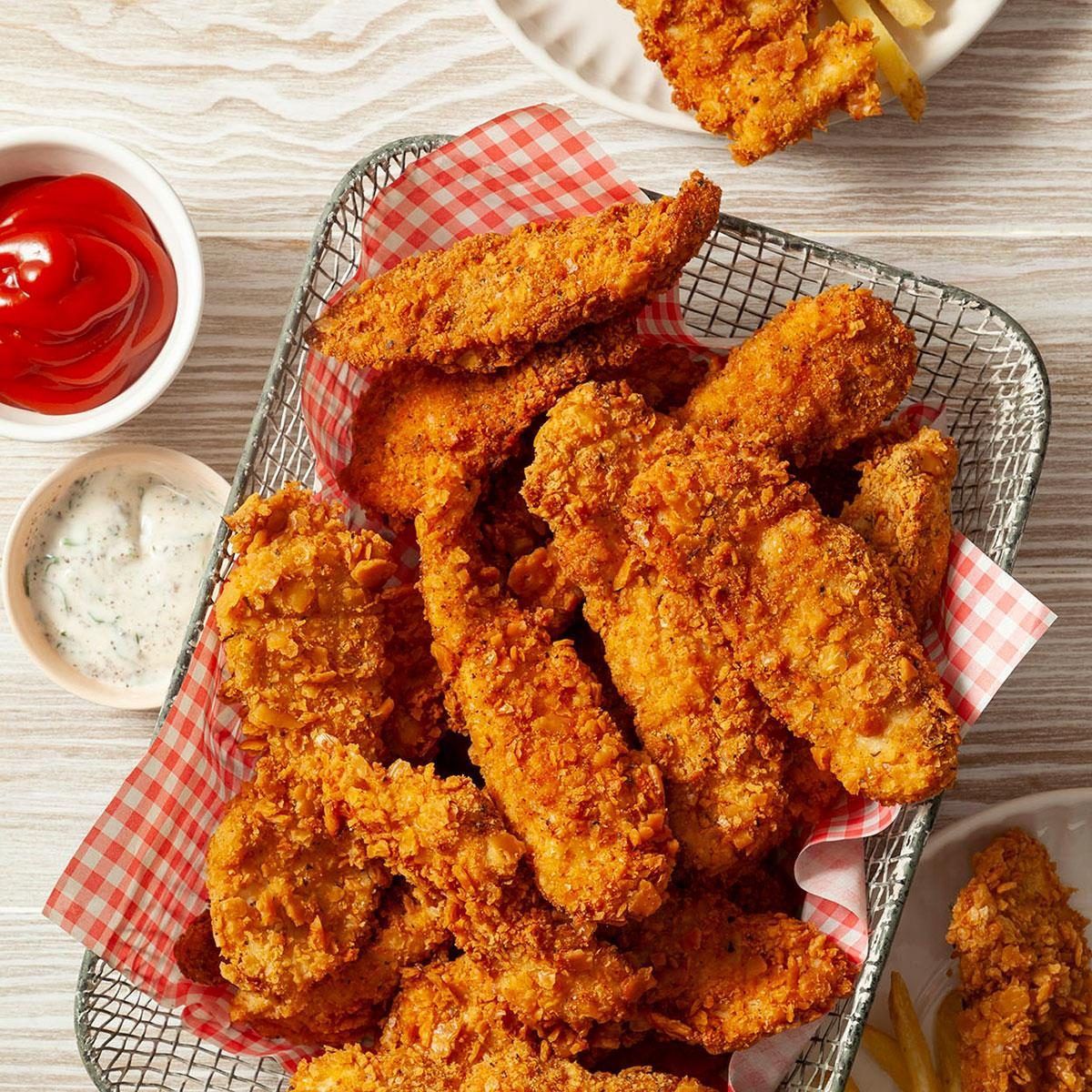 A wire basket lined with red and white checkered paper holds Chicken Zinger strips, fried in crispy seasoned breading. Dipping sauces and fries are served on the side.